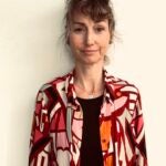 Photo of Christine. dark hair tied up in a messy bun, standing against a plain light-colored wall. She is wearing a brightly patterned button-up shirt in red, pink, orange, and white geometric shapes over a black top, a simple silver necklace with a small pendant.