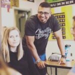 A smiling teacher leans on a desk next to a student.