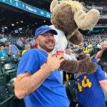 A baseball fan is standing in the bleachers at a Seattle Mariners game. He's throwing deuces and wearing a big smile, probably because he is standing next to the Mariners Moose Mascot.