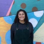 A woman in black overall and a black shirt smiles brightly in front of a colorfully painted wall.