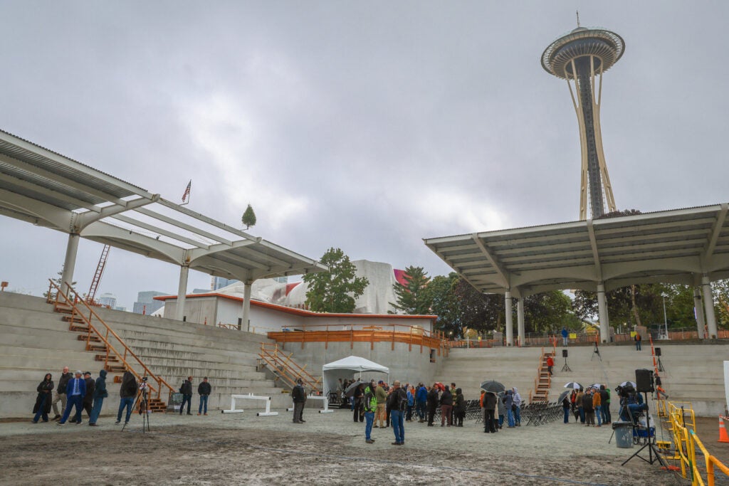 Wide view of Memorial Stadium under construction near the Seattle Space Needle. Concrete tiered seating surrounds an open gathering area where small groups of people stand near rows of chairs and a white event tent. Large steel canopy structures cover portions of the seating areas, and construction elements, including temporary stairs, railings, and equipment, are visible throughout the site. The Space Needle rises prominently in the background beneath an overcast sky.