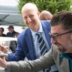 Superintendent Ben Shuldiner and another person in a gray jacket sign a white steel beam with black markers during a Memorial Stadium construction milestone celebration. The beam is covered with signatures and markings.