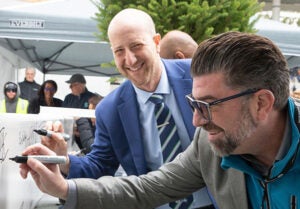 Superintendent Ben Shuldiner and another person in a gray jacket sign a white steel beam with black markers during a Memorial Stadium construction milestone celebration. The beam is covered with signatures and markings.