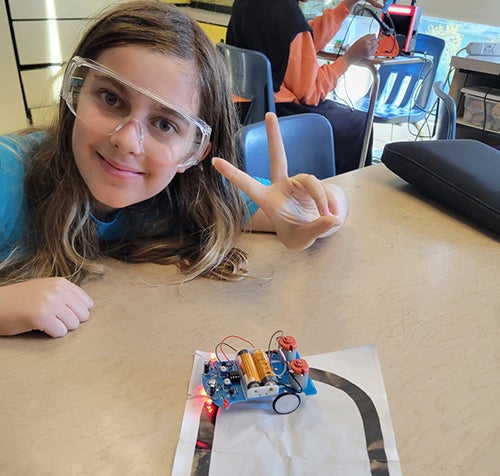 Student wearing safety glasses displays a small electronic robot built in class as it follows a track on a tabletop.