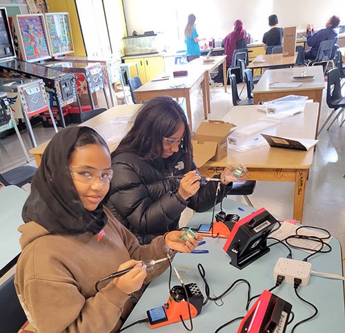 Two students wearing safety glasses assemble electronic kits in an ORCA K-8classroom. The students use soldering equipment, circuit boards, and instructional guides while learning electronics and engineering concepts. Pinball machines and additional students working on projects are visible in the background.