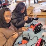 Two students wearing safety glasses assemble electronic kits in an ORCA K-8classroom. The students use soldering equipment, circuit boards, and instructional guides while learning electronics and engineering concepts. Pinball machines and additional students working on projects are visible in the background.