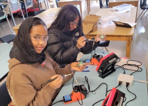 Two students wearing safety glasses assemble electronic kits in an ORCA K-8classroom. The students use soldering equipment, circuit boards, and instructional guides while learning electronics and engineering concepts. Pinball machines and additional students working on projects are visible in the background.