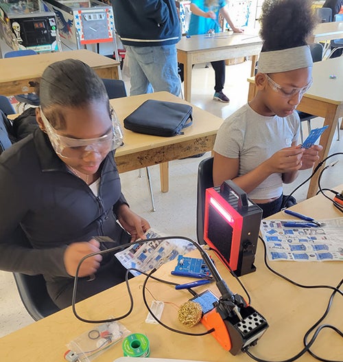 Two students wearing safety glasses work on electronics projects in a Orca K-8 classroom. Each student uses a soldering iron at a table equipped with soldering stations, hand tools, and electronic components. Additional students are working at tables in the background. Several pinball machines line one side of the classroom, highlighting a hands-on learning environment focused on electrical engineering and electronics.
