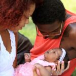 Two parents hold their baby in a grassy park.