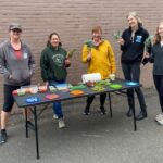 Five parent volunteers standing smiling around a table. They are holding leafy green vegetables and there are plates with small bites of vegetables to try.
