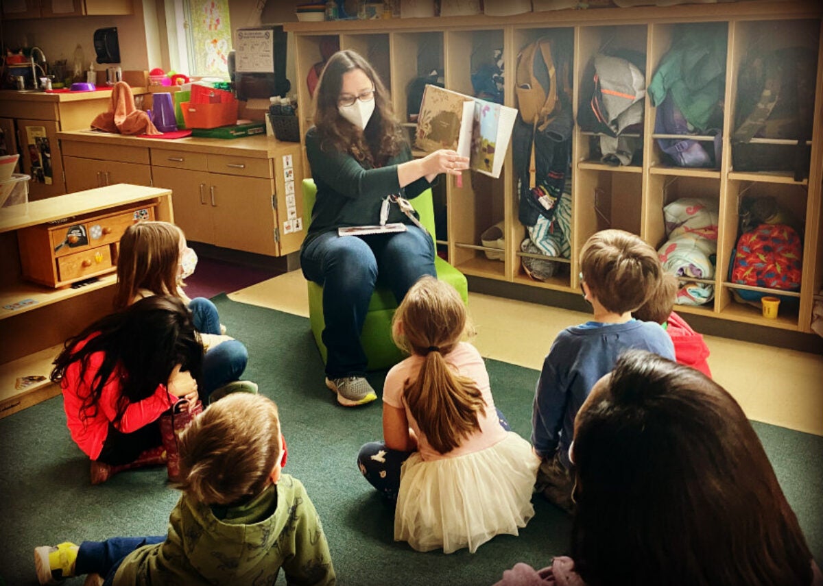 preschool educator reading a picture book to group of students on carpet