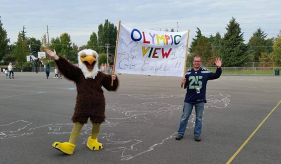 Mascot Beaky and Principal Bean on the black top holding an Olympic View banner