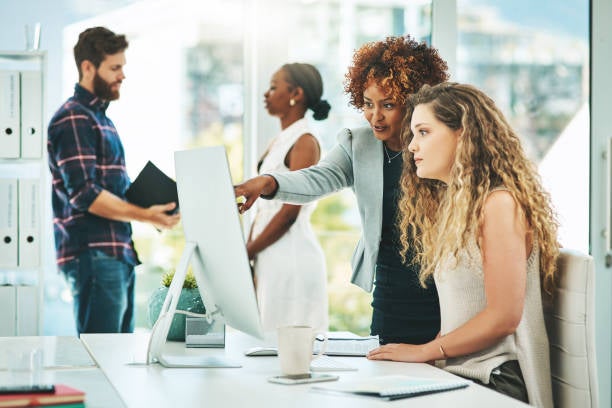 Shot of two women working together on a computer in an office with their colleagues in the background