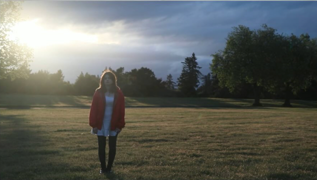 A person standing in the grass with clouds and sun glare behind them. Aurelia Film