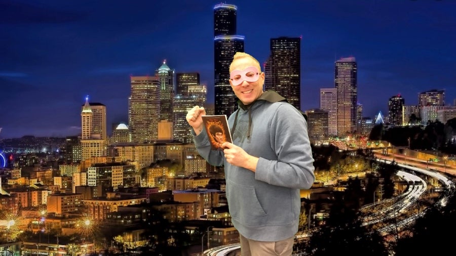 Mr. E stands in front of the Seattle skyline in a superhero mask with a book