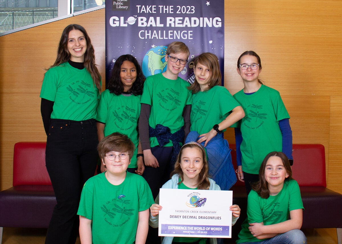 A group of students and a teacher gather for a photo in front of library Global Reading Challenge banner.